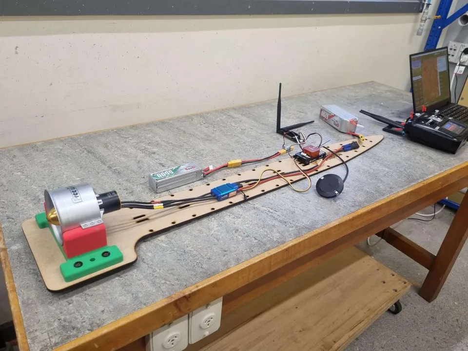 The avionics test bed: a long plywood board on a bench, laid out in the proportions of a fuselage. A ducted fan and motor sit in a printed mount at one end, feeding back through an ESC to a LiPo battery, with the flight controller, RF modem and antenna, and a round GPS puck spaced along the board. A laptop and transmitter sit at the far end.